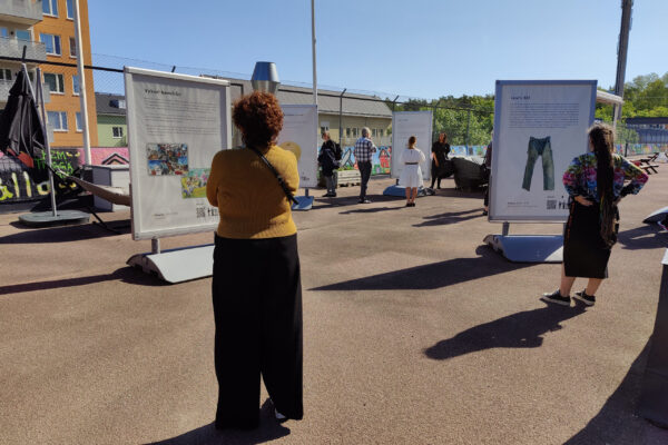 Nekoliko posjetilaca razgleda izložbu na otvorenom. U prvom planu je žena u žutoj majici