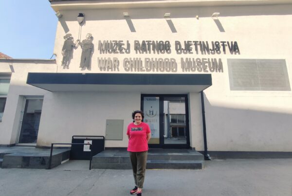 A young woman in reddish t-shirt stands outside the entrance to the War Childhood Museum. The sky is visible and clear behind her, and the WCM's logo is featured prominently